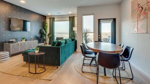 A modern living room with a green sofa, a round dining table with chairs, a TV mounted on the wall, and a large window with a view.