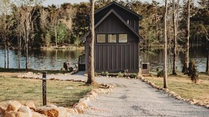 A small cabin with a gravel path leading to it, surrounded by trees and a body of water.