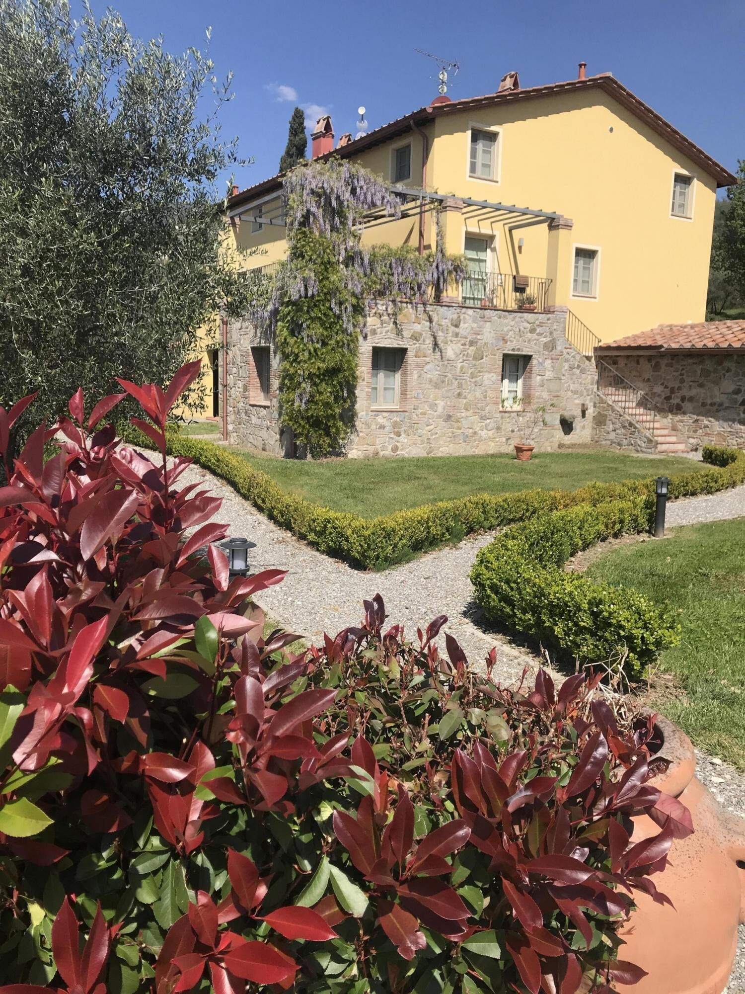 Un edificio amarillo con fachada de piedra, una escalera de piedra y un jardín bien cuidado con plantas de hojas rojas.