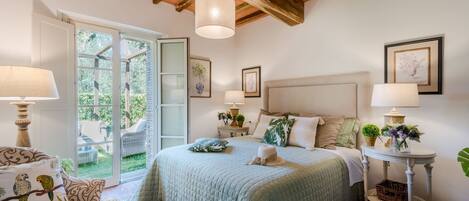 A bedroom with a large bed, two bedside tables with lamps, a chair, and a view of the garden through a glass door.