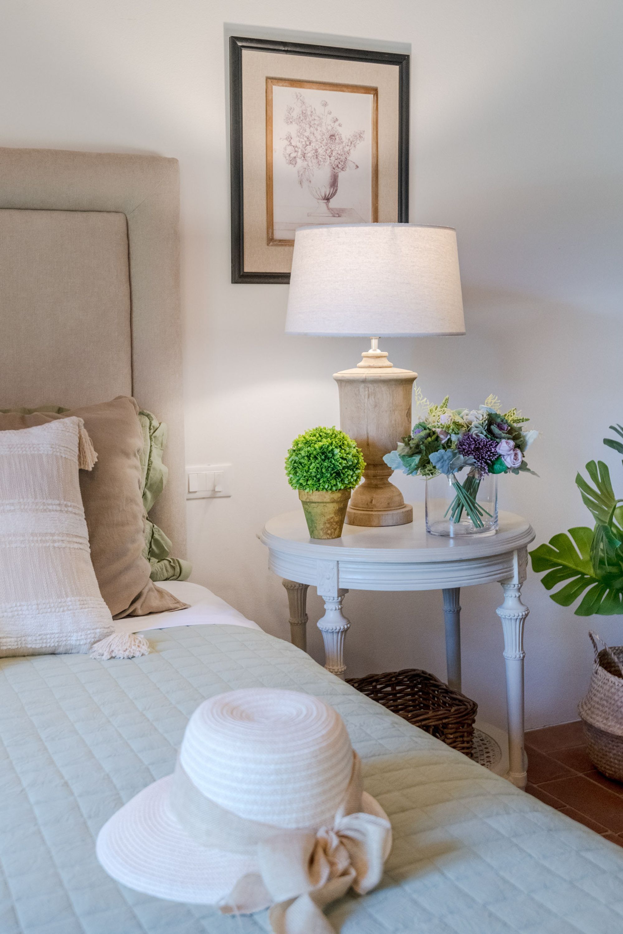 Un dormitorio ordenado con cama, una mesita de noche con lámpara, un cuadro enmarco y una pequeña planta en maceta.