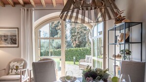 A dining area with a table set for two, a chair with a bird-themed pillow, and a bookshelf with decorative items. - Casa Pinocchio at Borghetto Farmhouse (Capannori)