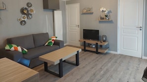 Una sala de estar con un sofá gris, una mesa de centro de madera, un televisor sobre un mueble bajo y un ventilador.