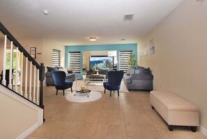A spacious living room with a staircase, a flat-screen TV, and a large window with blinds. - Family Getaway: Game Room, Themed Rooms & Private Yard Near Disney, BBQ!! (Kissimmee)