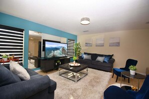 A modern living room with a flat-screen TV, a dark-colored sofa, a coffee table, and a blue armchair. - Family Getaway: Game Room, Themed Rooms & Private Yard Near Disney, BBQ!! (Kissimmee)