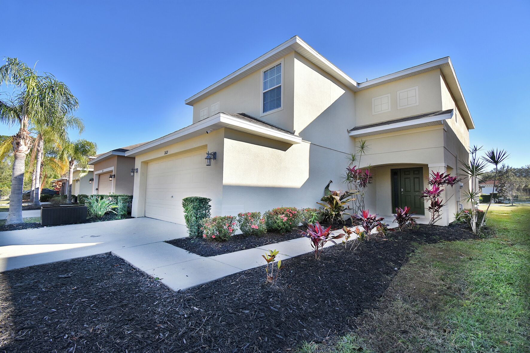 A two-story residential house with a garage, landscaped garden, and palm trees.
