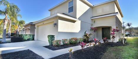 A two-story residential house with a garage, landscaped garden, and palm trees.
