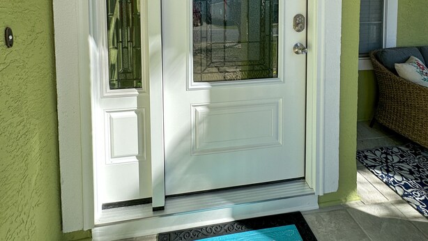 Une porte d’entrée blanche avec un panneau de verre, un paillasson sur lequel est inscrit « Sandvilla Beach House », et une chaise en osier avec des coussins.