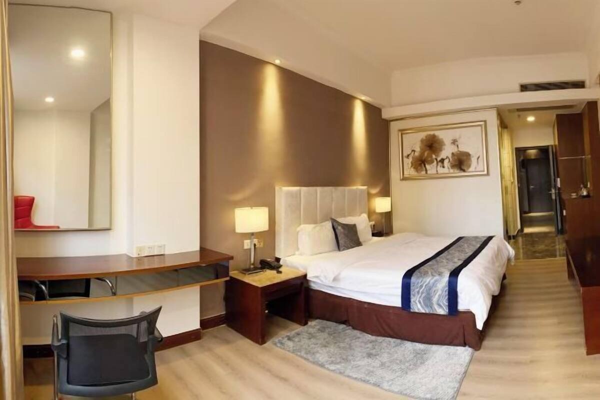 A hotel room with a bed, desk, chair, bedside lamps, and a painting on the wall.