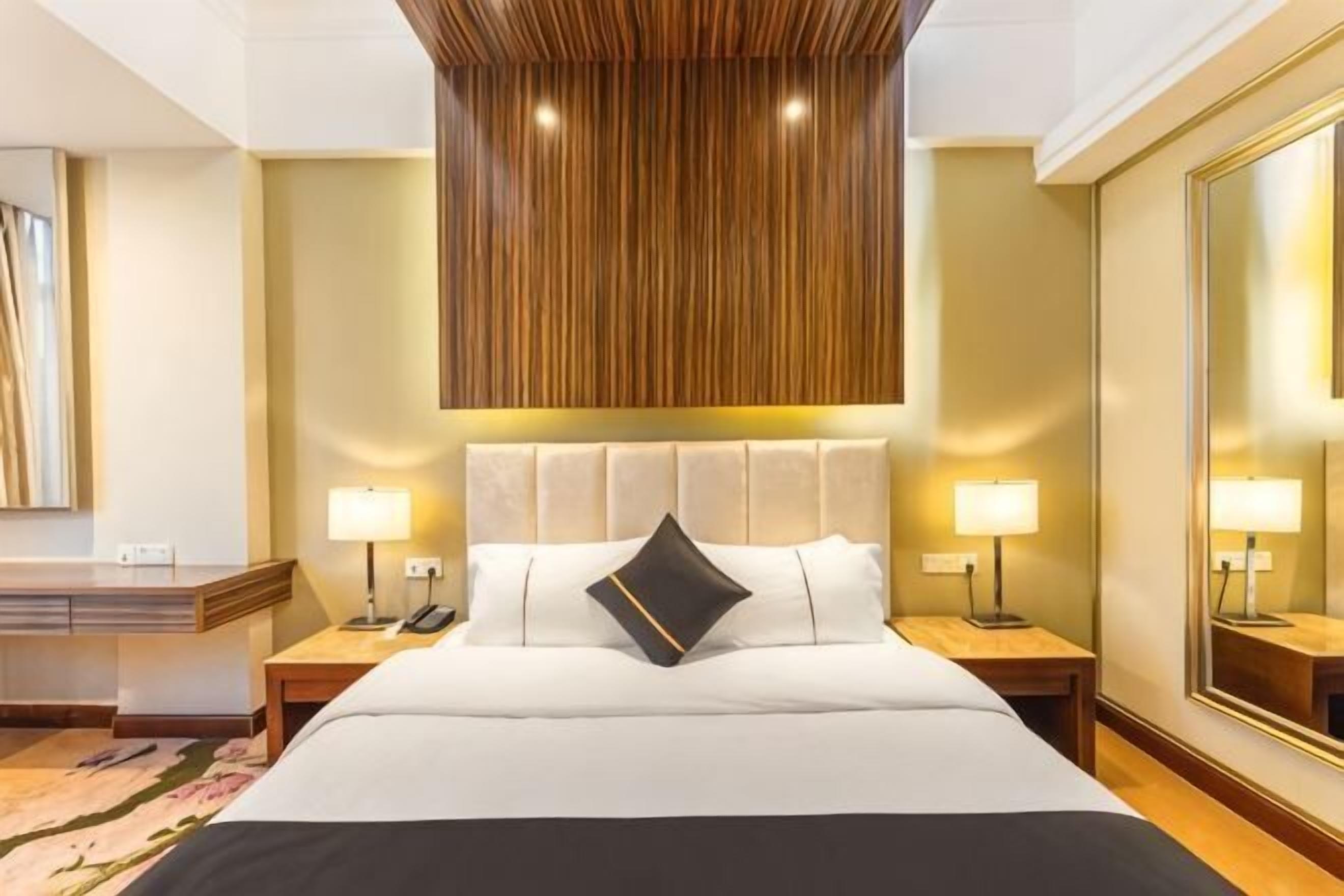 A hotel room with a large bed, bedside tables with lamps, a wooden ceiling panel, and a large mirror on the wall.