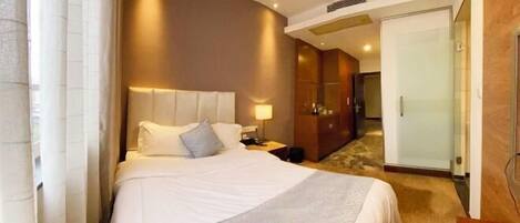 A hotel room with a bed, bedside table, lamp, wardrobe, and a door leading to a hallway.