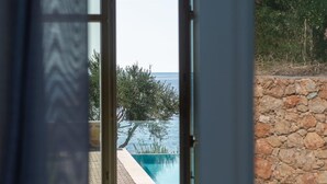A view through a window of a swimming pool with a stone wall and greenery in the background. - Villa I With Private Pool (West Mani)