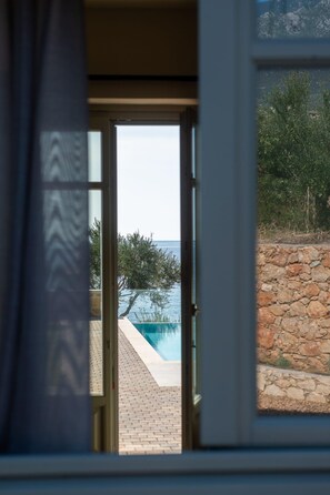 A view through a window of a swimming pool with a stone wall and greenery in the background. - Villa I With Private Pool (West Mani)
