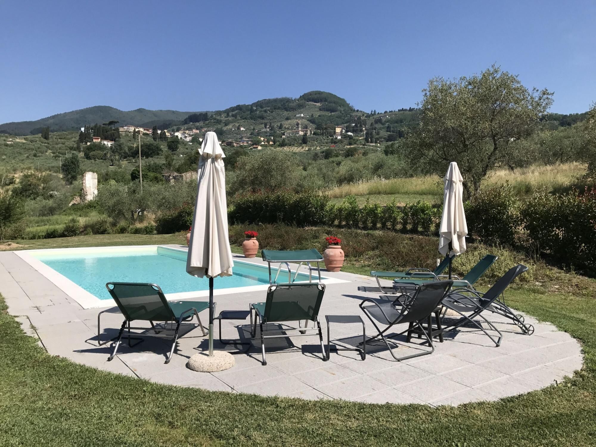Un espace piscine agrémenté de chaises longues et de parasols, entouré d’un jardin aménagé et offrant une vue sur une ville perchée sur une colline.
