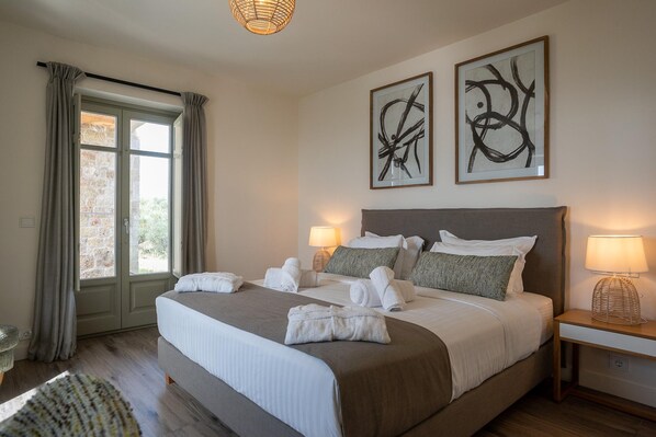 A hotel room with a large bed, two bedside lamps, a small table, and a view through a glass door. - Villa Stoupa Exclusivity (West Mani)