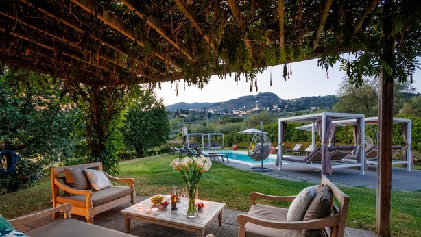 Un patio al aire libre con mobiliario de madera, una mesa con flores y una piscina al fondo.