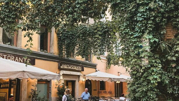 An outdoor café with tables and chairs, a bicycle, and a building with a sign reading "all'arcade di S. Calisto".