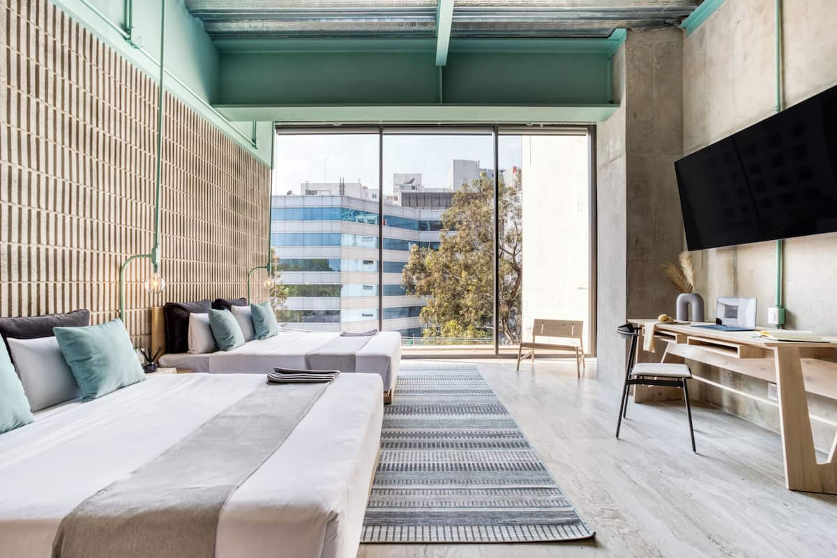 A modern hotel room with a large bed, a desk, and a view of the city through large windows.