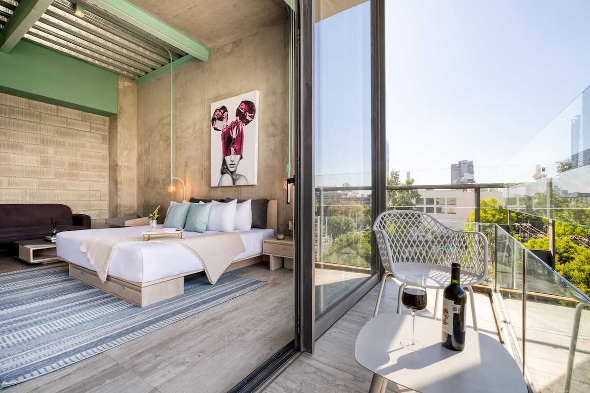 A modern bedroom with a large bed, a balcony with a glass railing, and a view of the city.