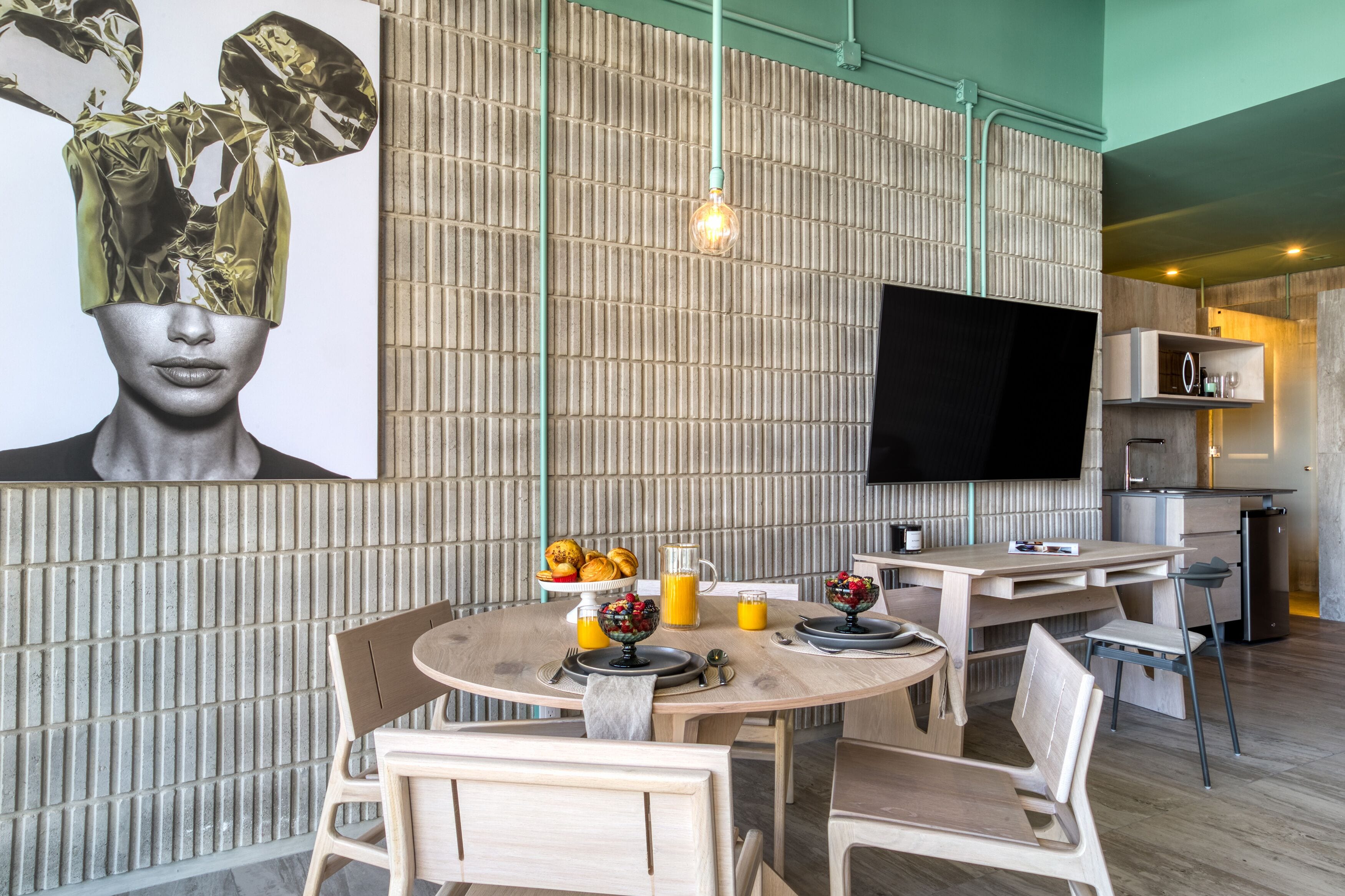 Un espace repas moderne avec une table ronde en bois, dressée pour deux personnes, un grand téléviseur à écran plat fixé au mur, et un coin cuisine visible au fond.