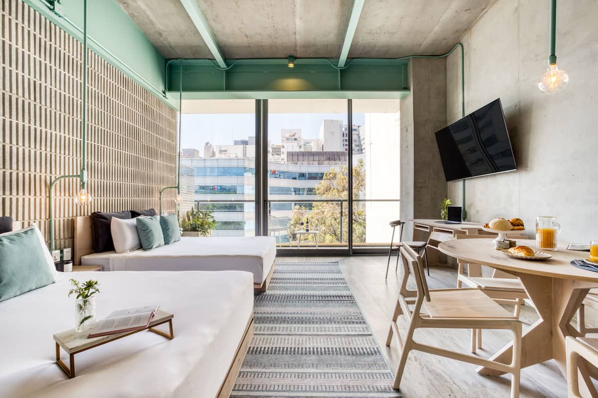 A modern hotel room with a large window, a bed, a dining table with breakfast, and a TV mounted on the wall.