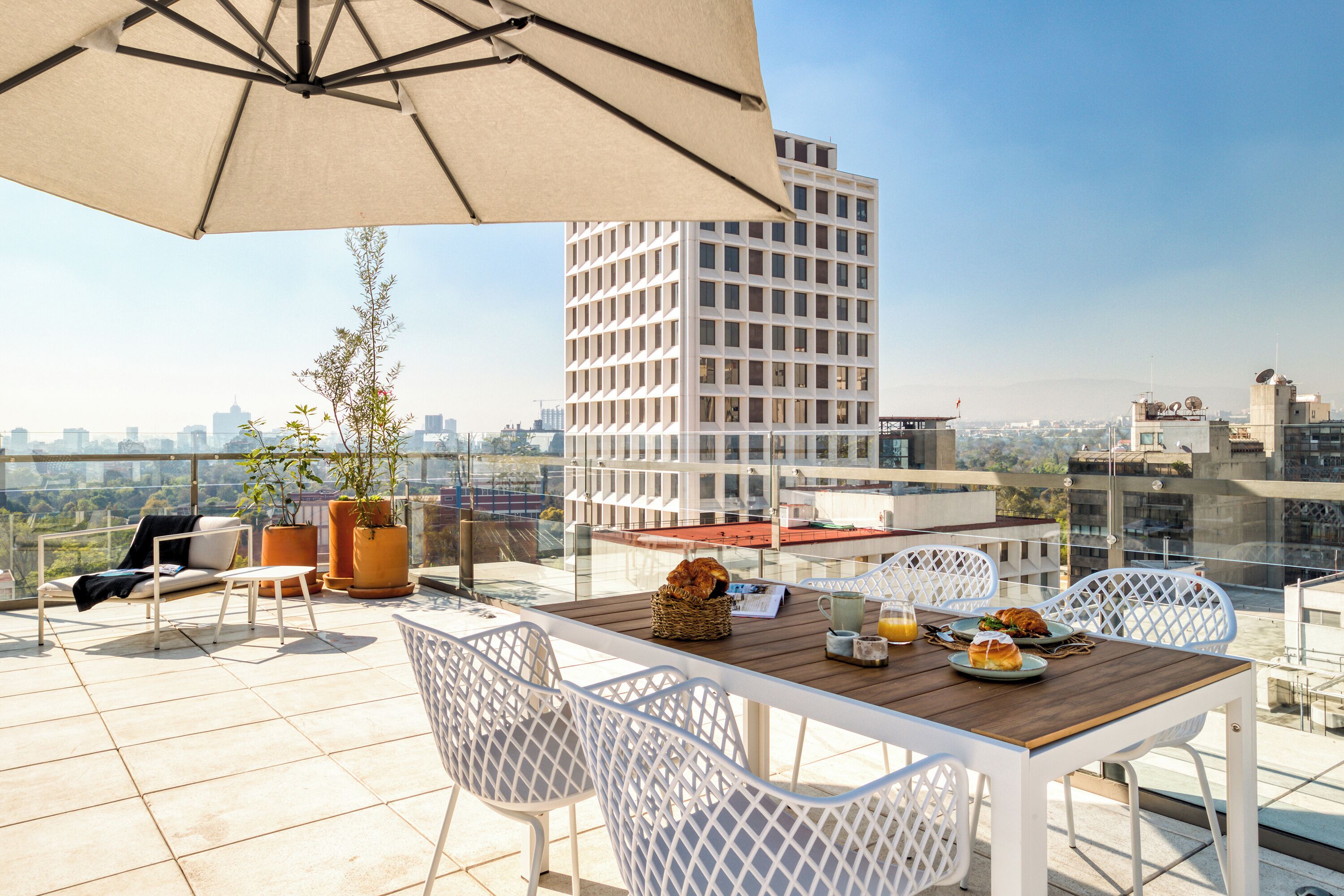 A rooftop terrace with a table set for breakfast, white chairs, a large umbrella, and a city view.
