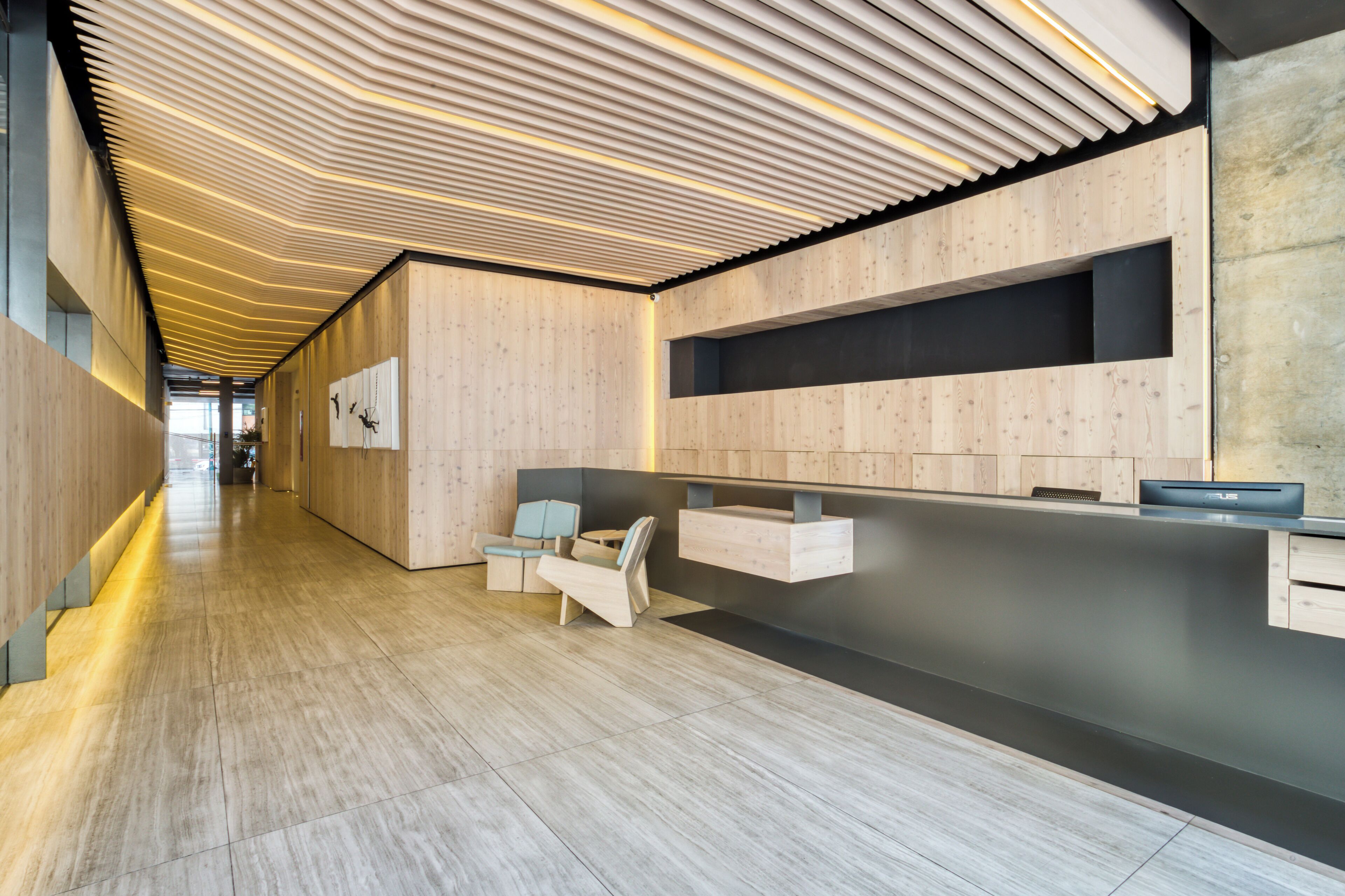 A modern reception area with a wooden reception desk, seating area, and a long corridor with a wooden floor.