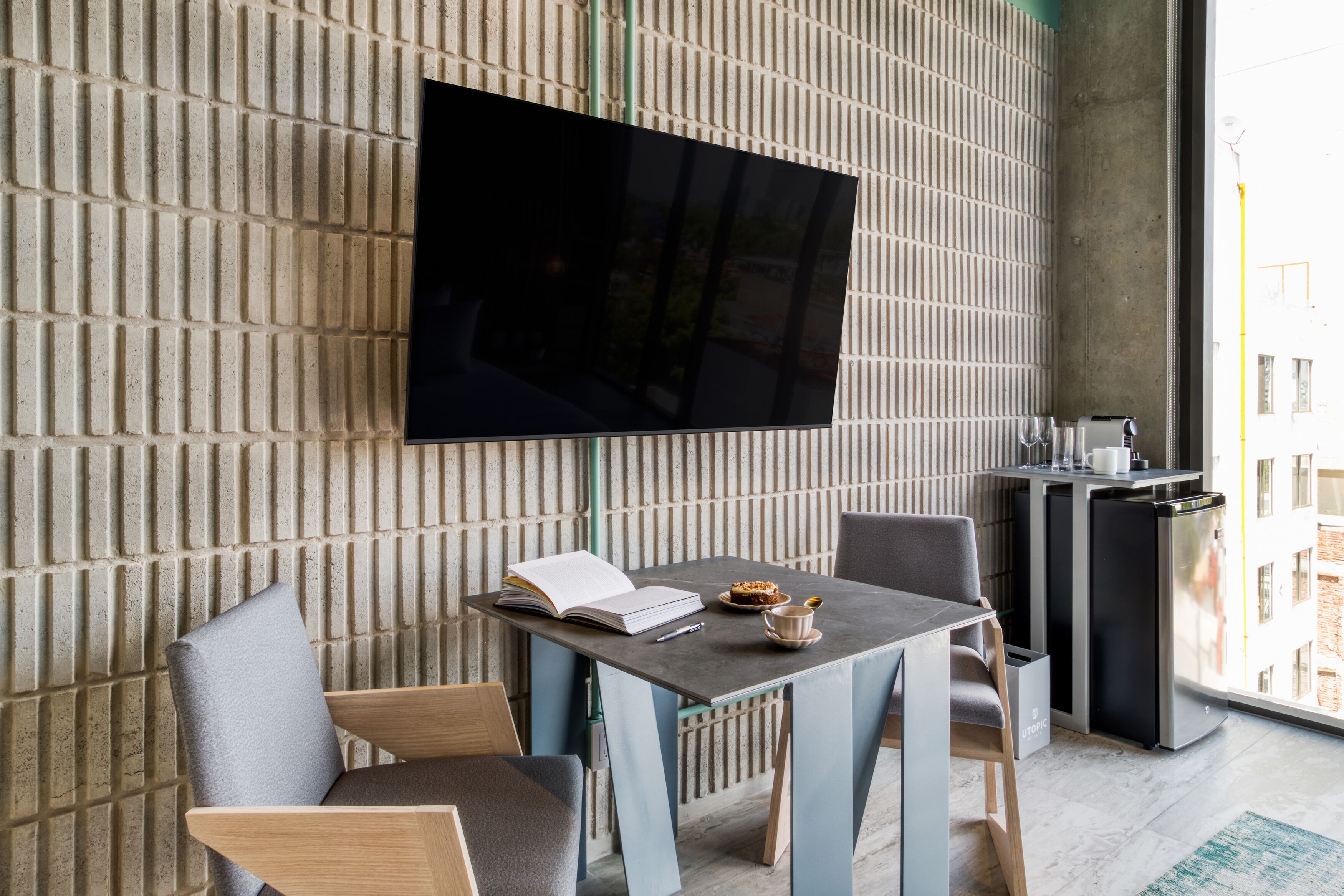 A modern room with a flat-screen TV mounted on a textured wall, a small table with a book and a cup, and a compact kitchenette with a coffee machine.