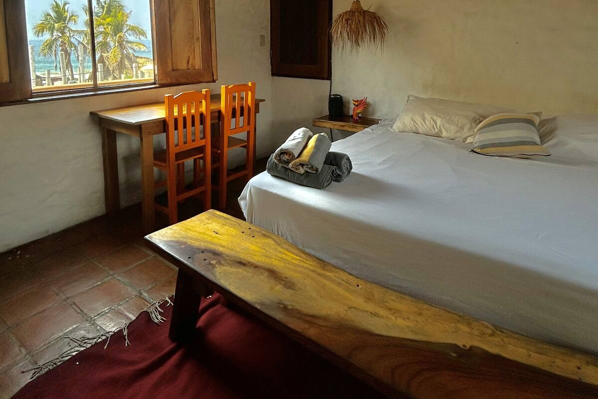 A room with a bed, a desk, a chair, a ceiling fan, and a window with a view of palm trees.