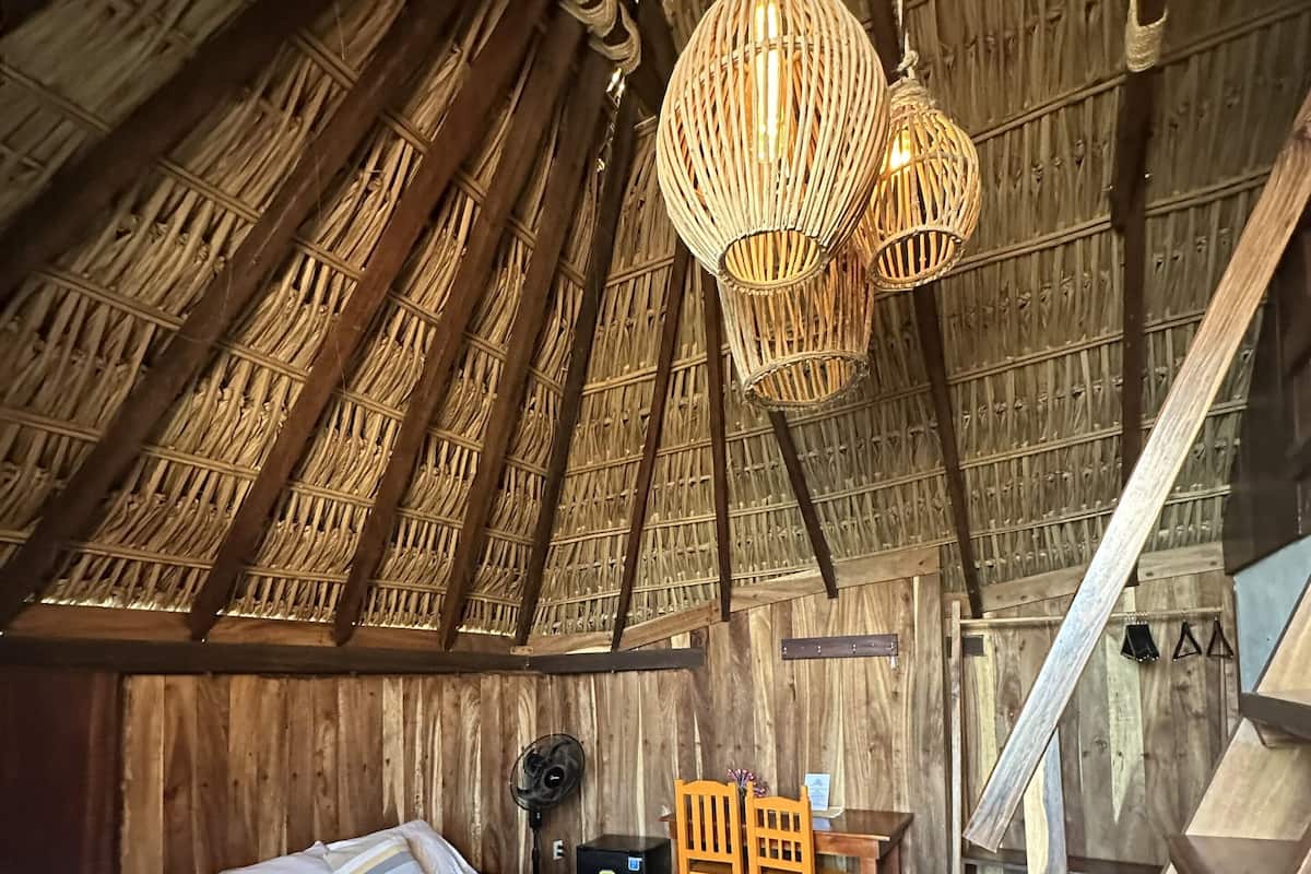A rustic room with a bed, wooden walls, a woven ceiling light, and a small table with chairs.