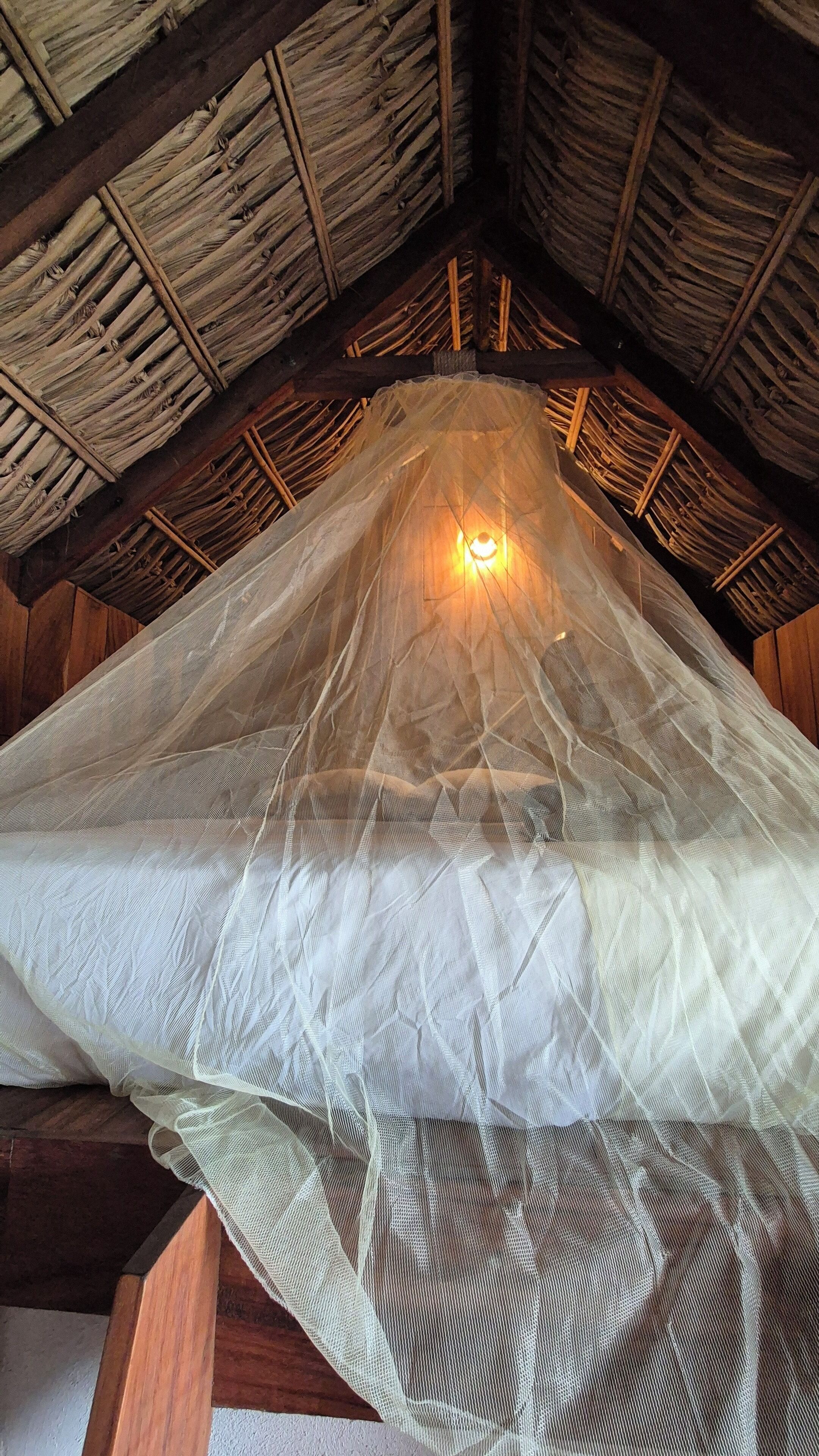 Ein Zimmer mit Strohdach und einem weißen, halbtransparenten Baldachin über dem Bett.