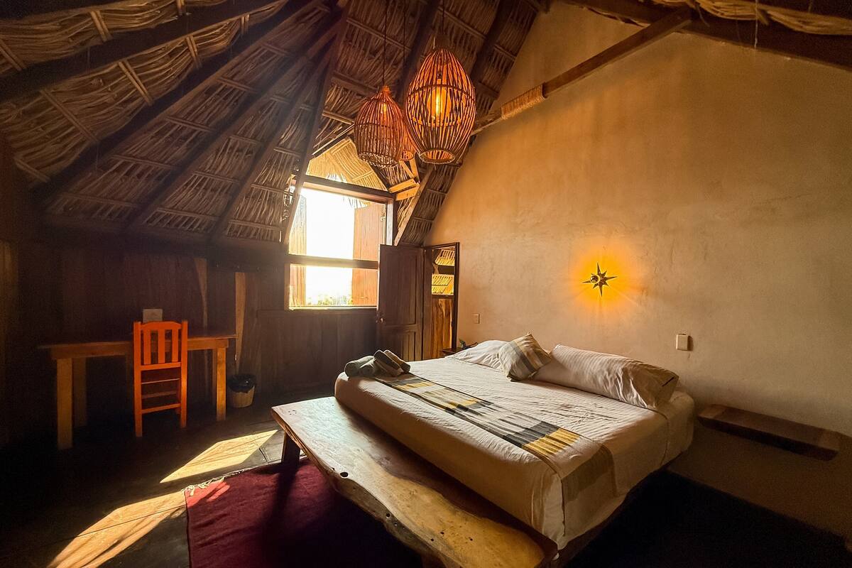 A rustic room with a bed, a wooden chair, a table, and a thatched roof.