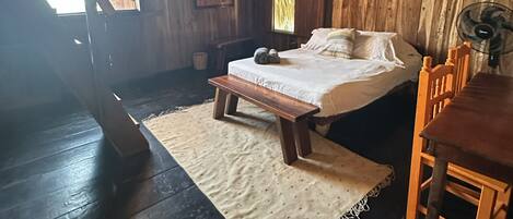 Una habitación rústica con techo de madera, una cama con almohadas, un banco, una mesa y un ventilador.