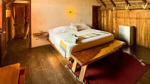 A rustic room with a sloped ceiling, a bed with a wooden frame, a small bedside table, a nightstand, a wall-mounted light, and a wooden door. - Casa Playa del Amor (San Pedro Pochutla)
