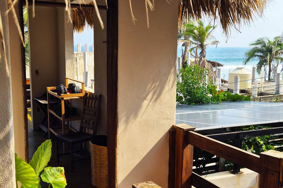 A thatched-roof balcony with a view of the sea, a wooden table, chairs, and a wicker basket.