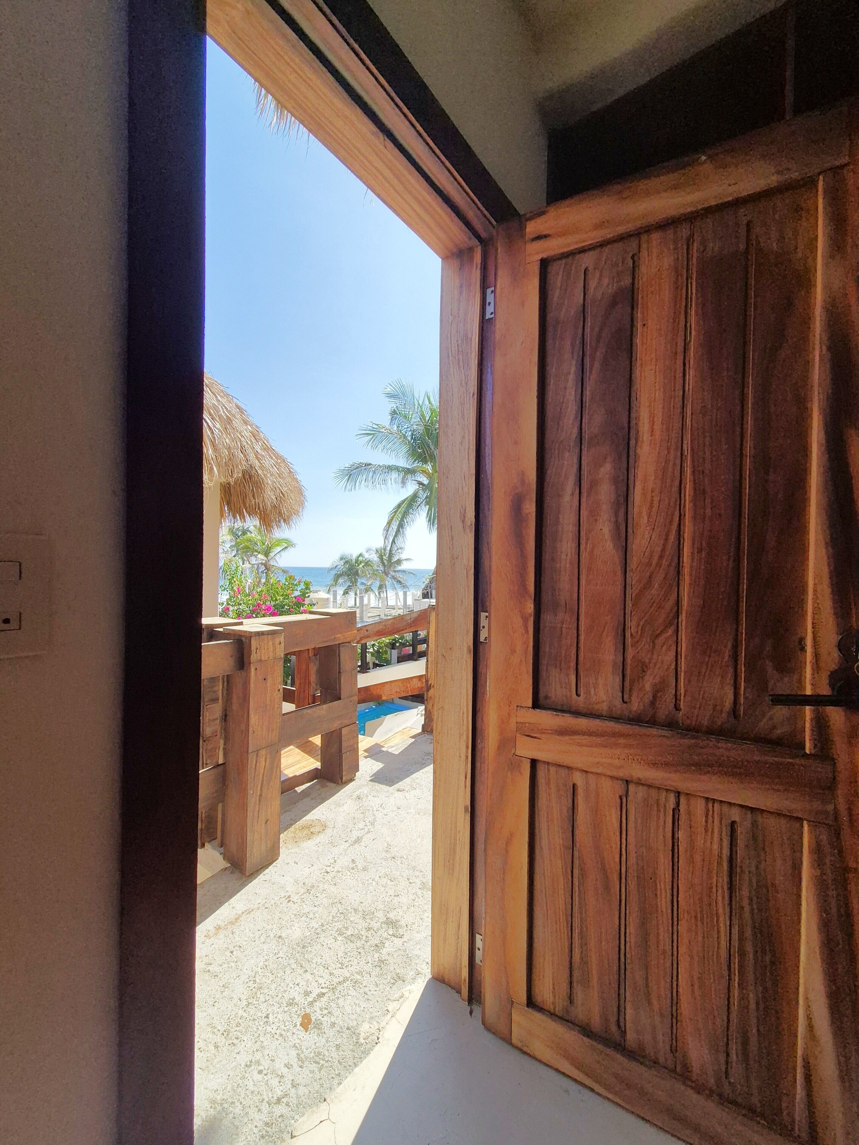 A wooden door leading to a beach view with a thatched roof structure and palm trees.