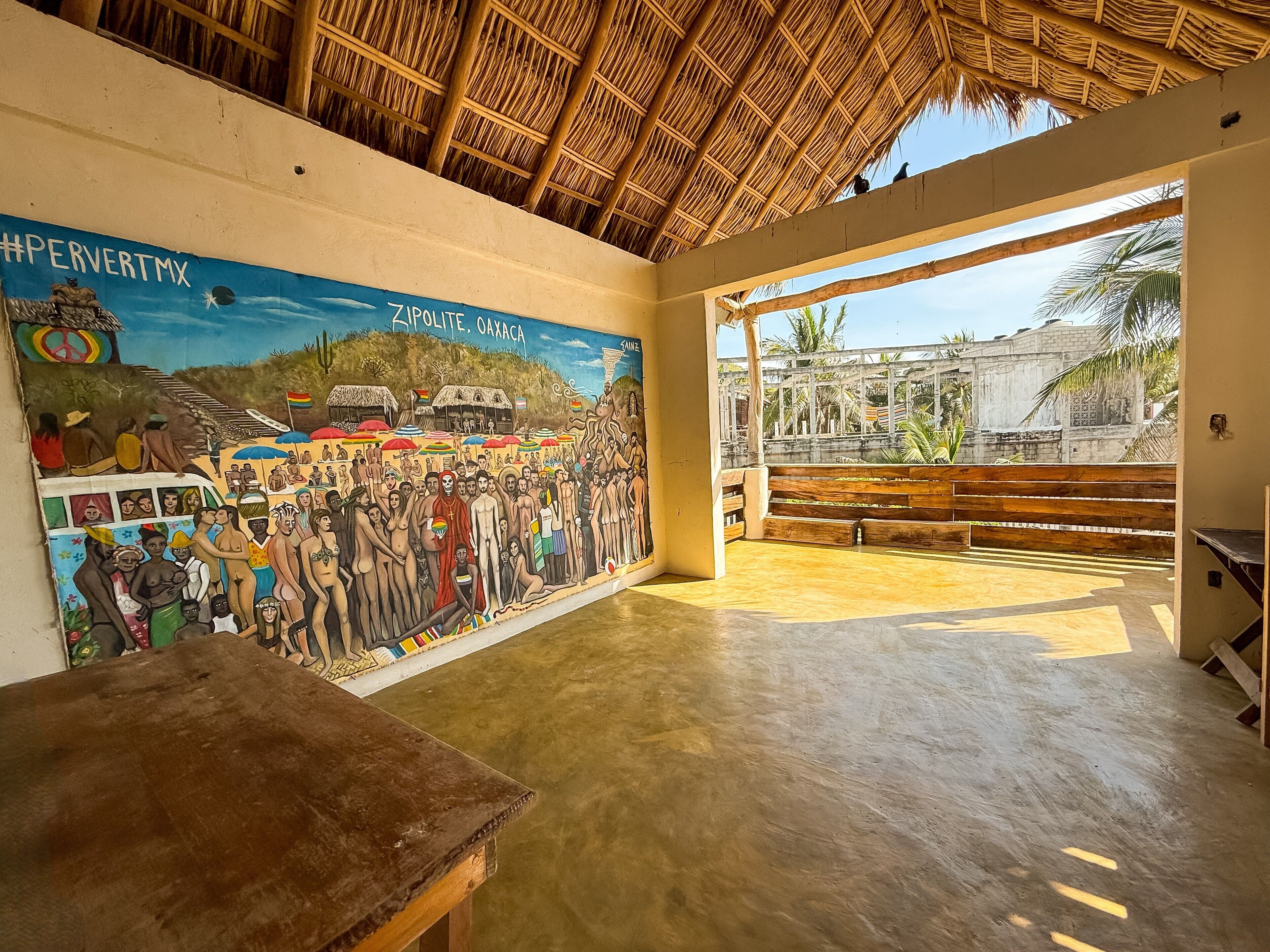 Un espacio interior con un mural que representa una escena histórica, techo de paja y vistas a palmeras fuera.