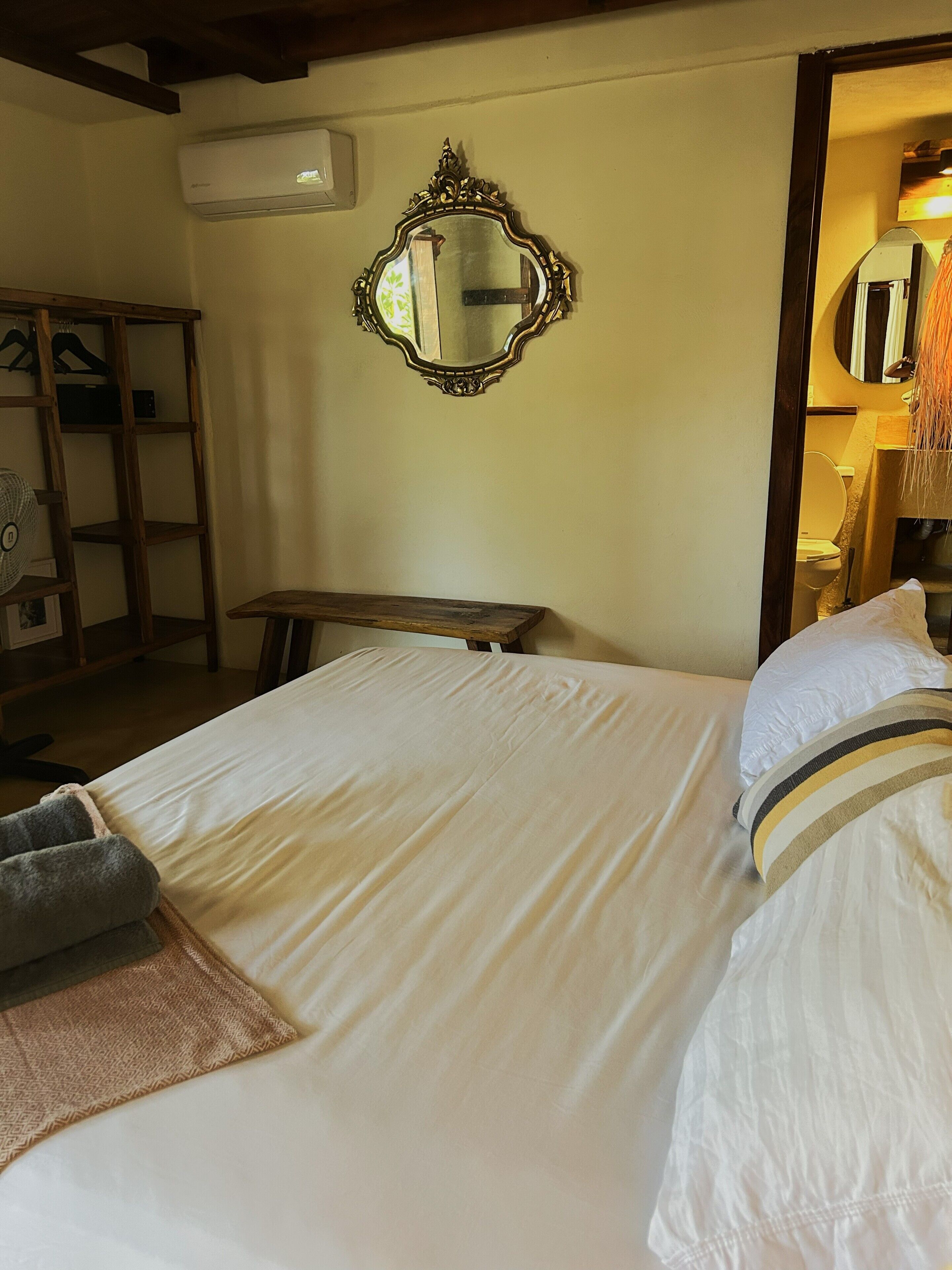 A room with a bed, a wooden bench, a mirror, and a wall-mounted air conditioner.