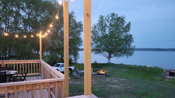 A wooden deck with string lights, outdoor furniture, and a view of a lake. - Modern Retreat on Lake Superior, close to the Snowmobile trails! (Baraga)