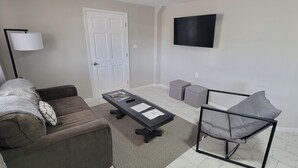 A living room with a brown sofa, a coffee table with magazines and a remote, a grey armchair, a floor lamp, and a TV on the wall. - Modern Retreat on Lake Superior, close to the Snowmobile trails! (Baraga)