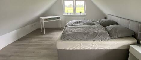 Ein Schlafzimmer mit schrägen Wänden, einem Bett mit grauer Bettwäsche, einem kleinen weißen Nachttisch und einem Fenster mit Blick auf ein grünes Feld.