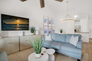 A modern living room with a light blue sofa, a flat-screen TV mounted on the wall, a chandelier, and a kitchen area in the background.
