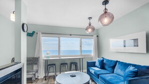 Un salón con un sofá azul, una mesa de centro redonda y un ventanal con vista al mar. Se observan tres taburetes y una mesita con una lámpara.