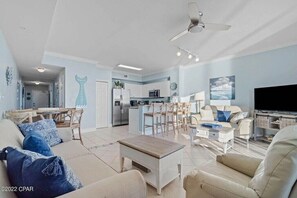 Una sala moderna con sofá, mesa de centro y televisor. Cuenta con una zona de cocina con refrigerador, microondas y mesa de comedor. La habitación tiene ventilador de techo e iluminación empotrada.