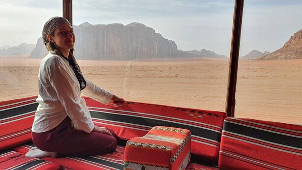 A person sitting on a red and black striped carpet inside a room with large windows overlooking a desert landscape with mountains. - Wadi Rum Oasis (Wadi Rum)