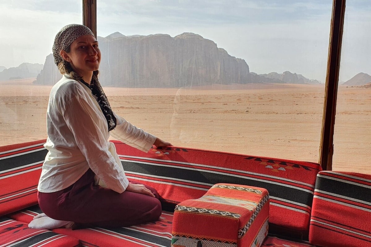 A person sitting on a red and black striped carpet inside a room with large windows overlooking a desert landscape with mountains.