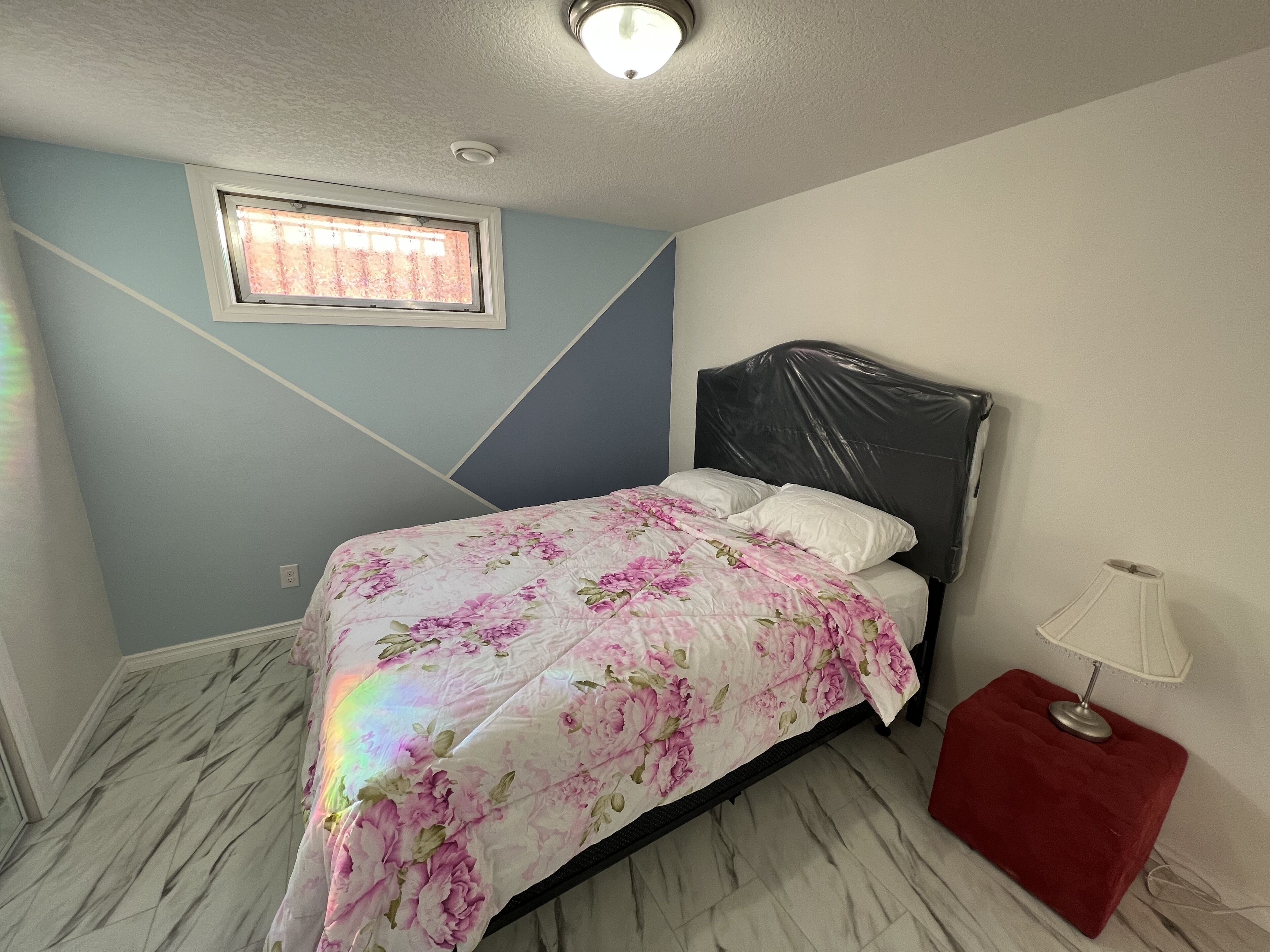 Ein Schlafzimmer mit einem Bett, einem Fenster, einer Lampe und einem roten Hocker.
