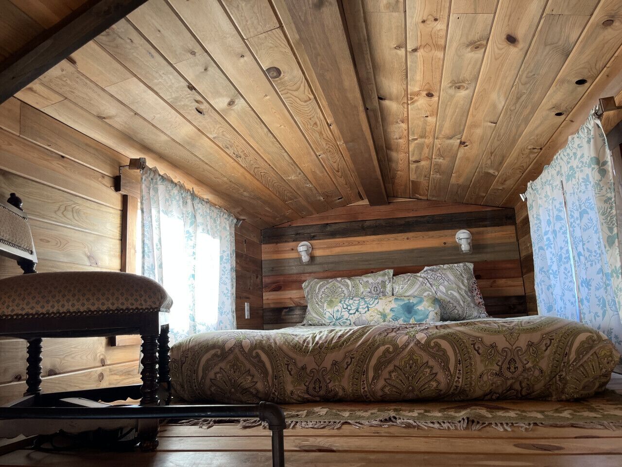 Ein Schlafzimmer einer Holzhütte mit Bett, gemusterten Textilien, einer Bank und einem Fenster mit Vorhängen.
