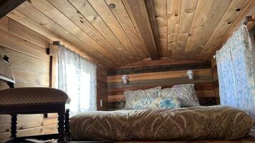 Ein Schlafzimmer einer Holzhütte mit Bett, gemusterten Textilien, einer Bank und einem Fenster mit Vorhängen.