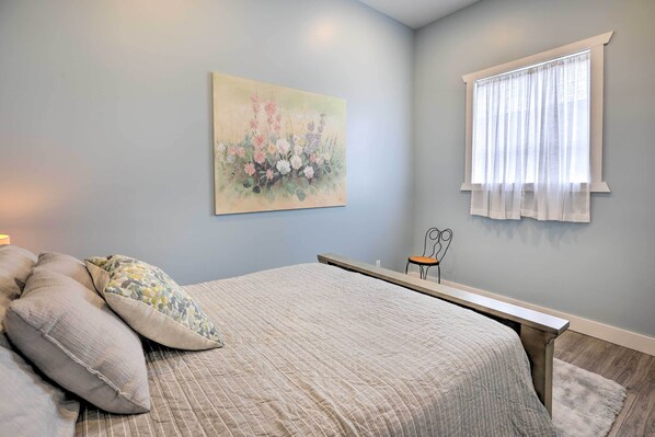 Een slaapkamer met een bed, kussens, een nachtkastje, een raam met gordijnen en een schilderij aan de muur.